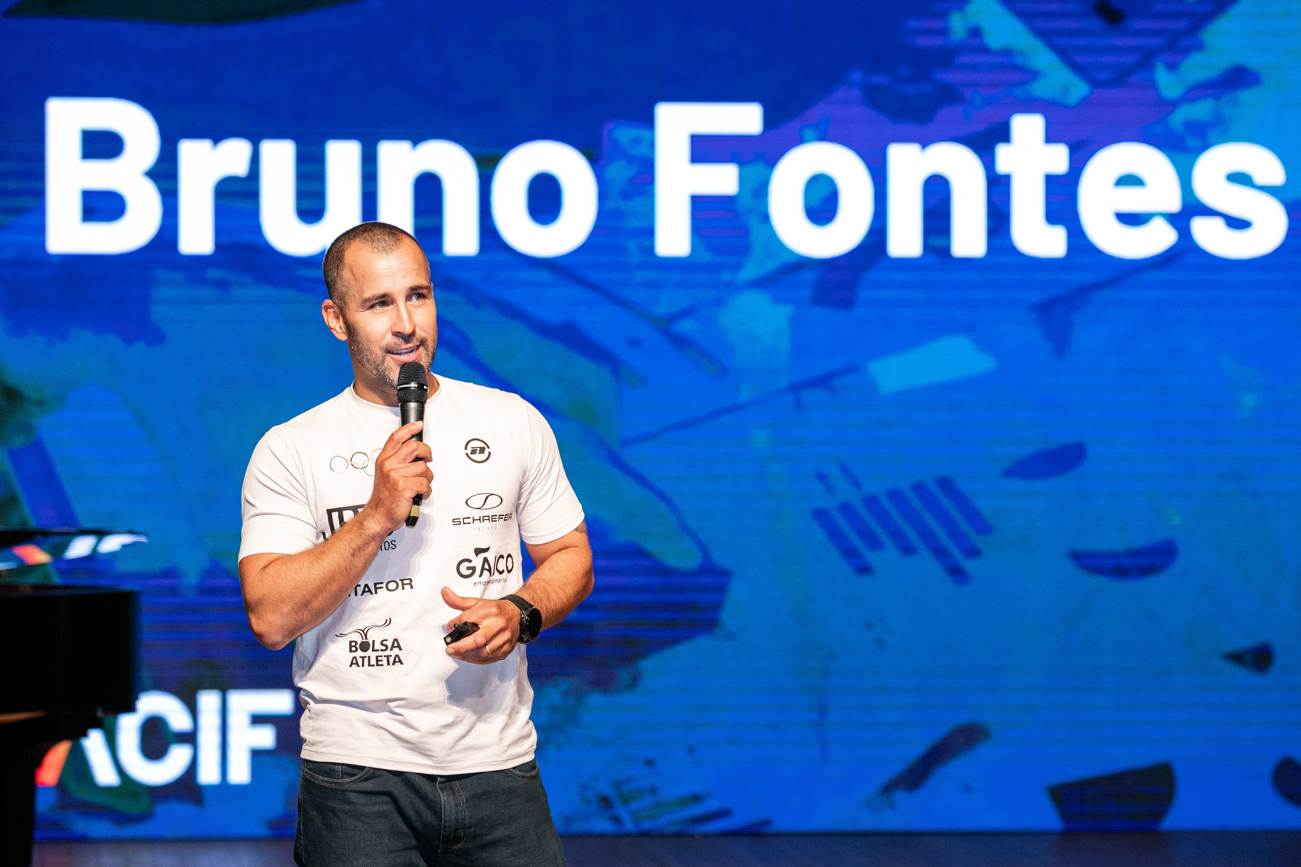 Bruno Fontes no palco com seu nome projetado na tela durante palestra corporativa
